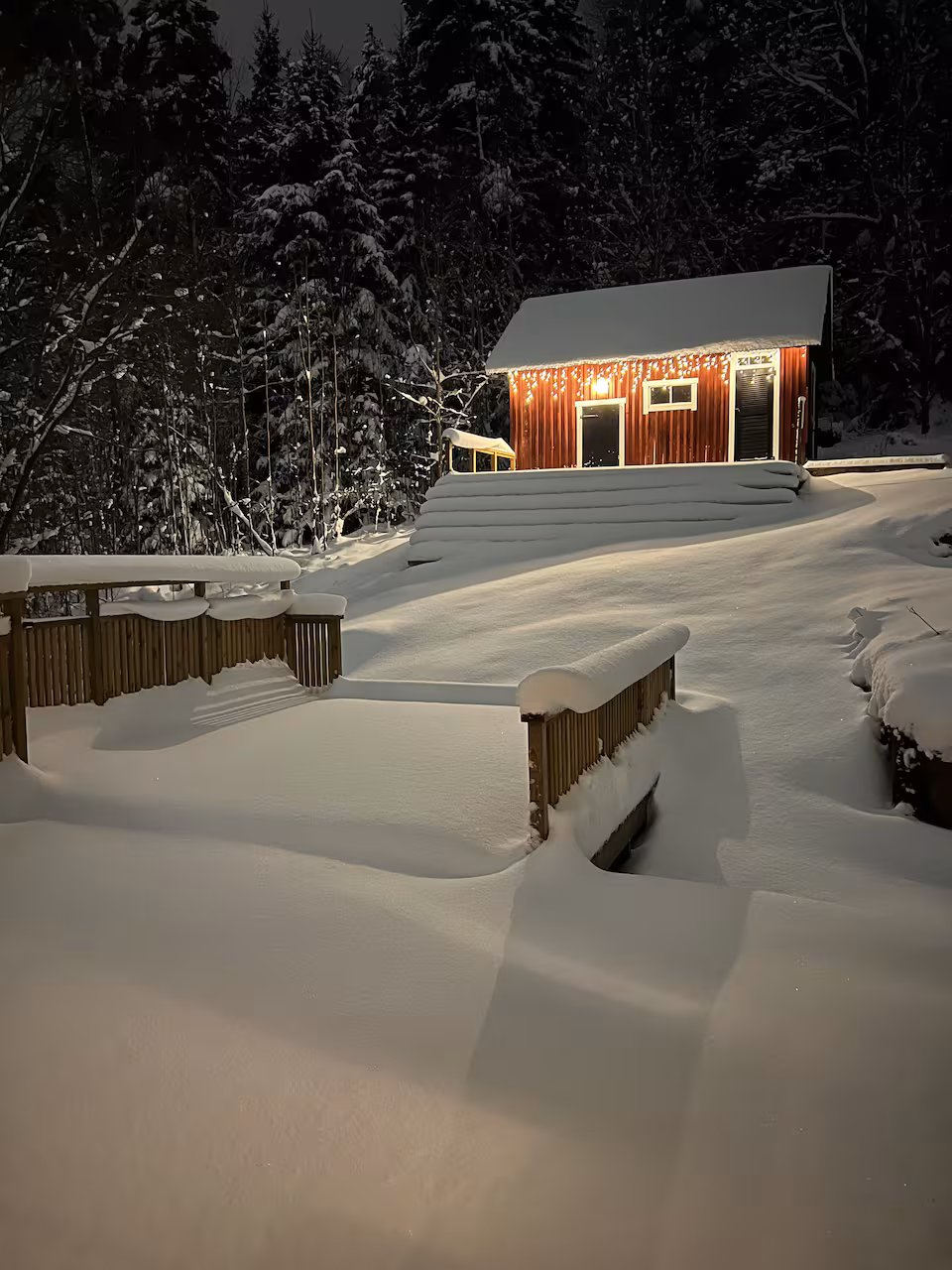 Röd stuga i djup snö en vinterkväll, upplyst bland granar