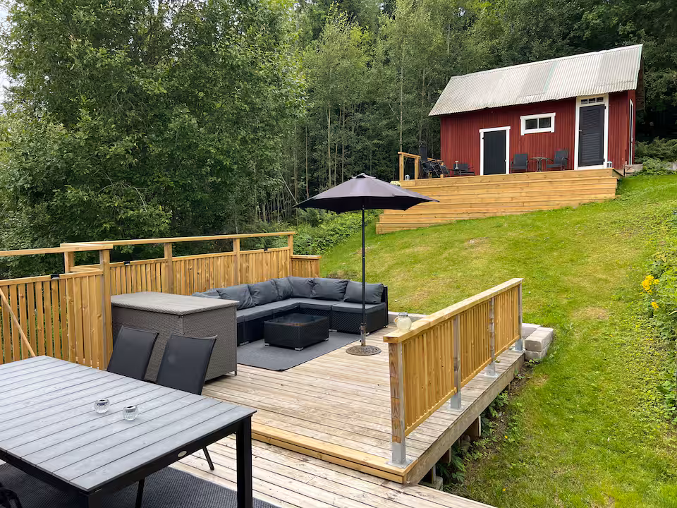 Trädäck med loungemöbler och parasoll, gräsmatta och en röd bod i sommargrönska