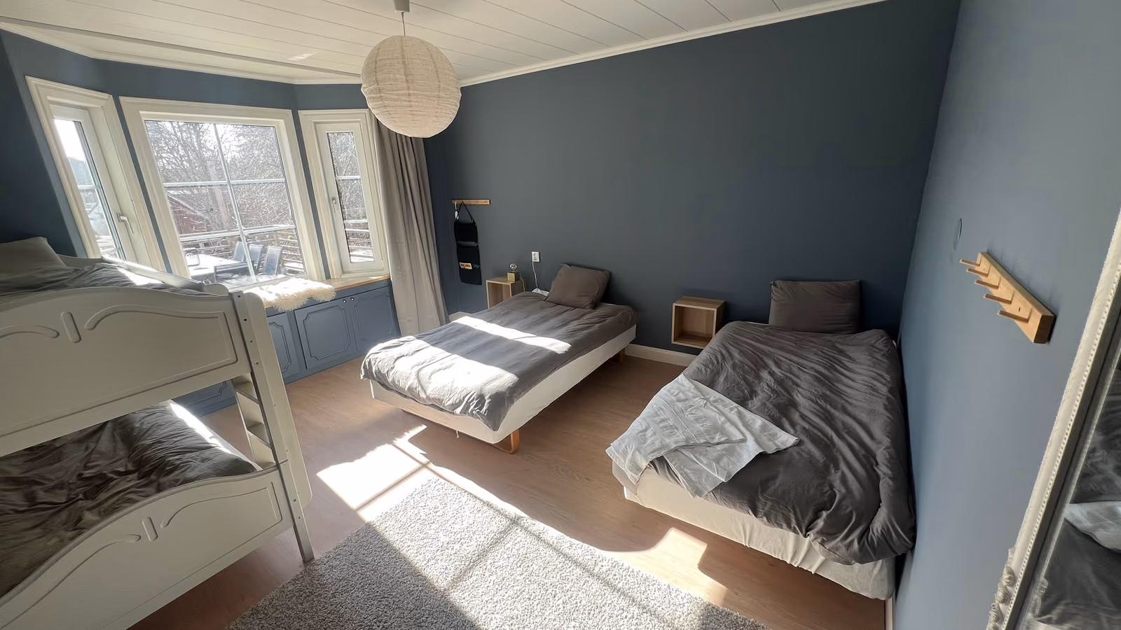 Soligt sovrum med blå väggar, våningssäng, två sängar och burspråk mot terrassen